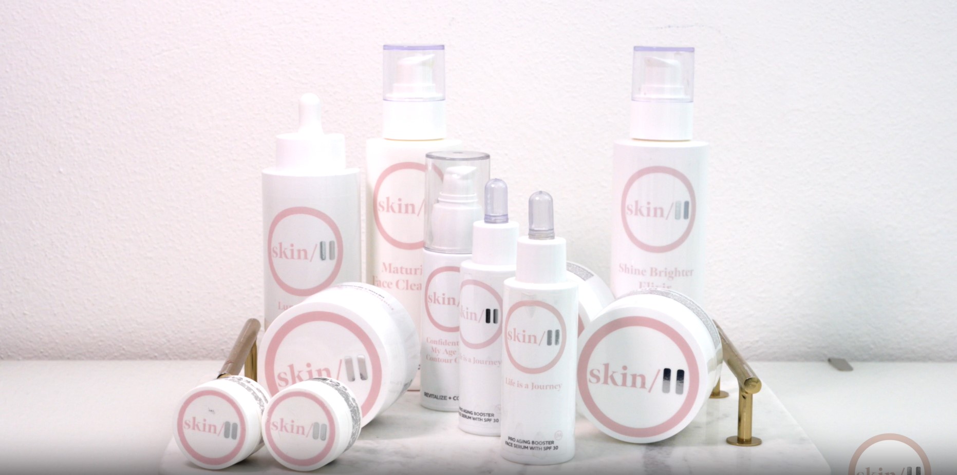 Diversi flaconi, vasetti e contagocce bianchi di prodotti per la cura della pelle Skinpause con il marchio della pelle rosa e grigio sono disposti ordinatamente su uno scaffale bianco su uno sfondo chiaro. I prodotti variano per dimensione e forma.